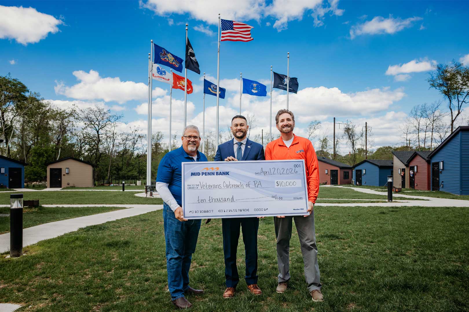 Three people hold a $10,000 donation check for Veterans Outreach of Pennsylvania in front of flagpoles and veteran housing cabins.